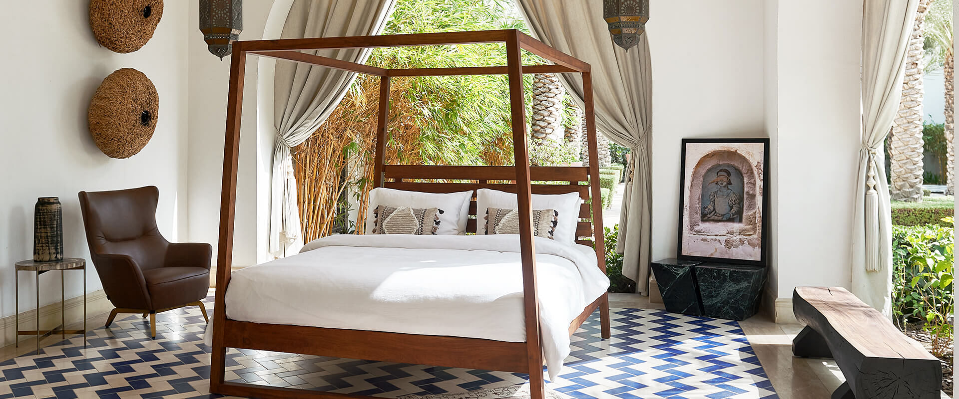 A four poster bed stands by the windows on a blue-and-white tiled floor. A leather armchair,  wooden bench, and wall art are positioned nearby.