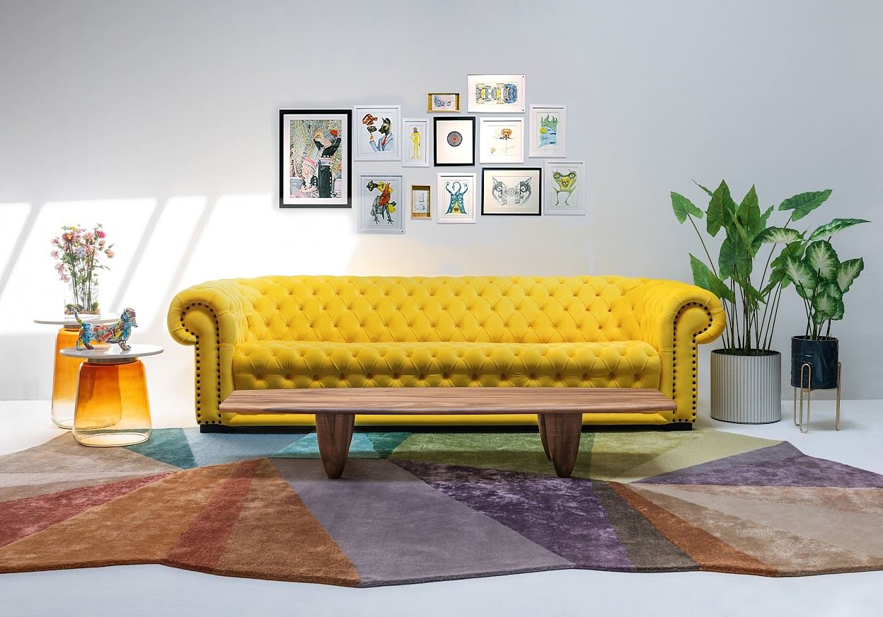 A yellow sofa is paired with a multi-coloured rug, glass tables, vibrant artwork, and plants