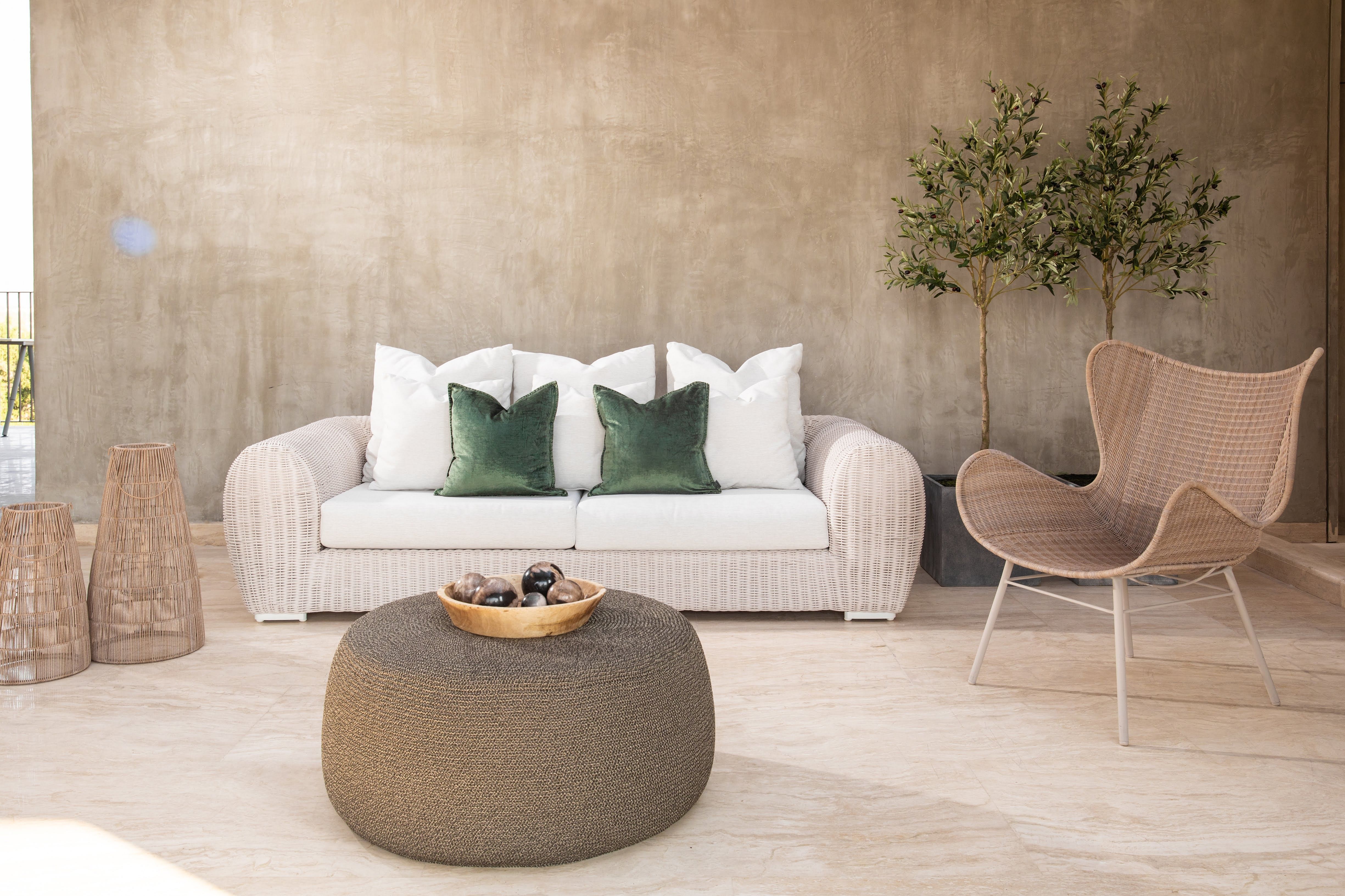 A white two-seater sofa stands against a taupe wall. A rattan armchair, coffee table, accessories and miniature olive trees stand nearby.
