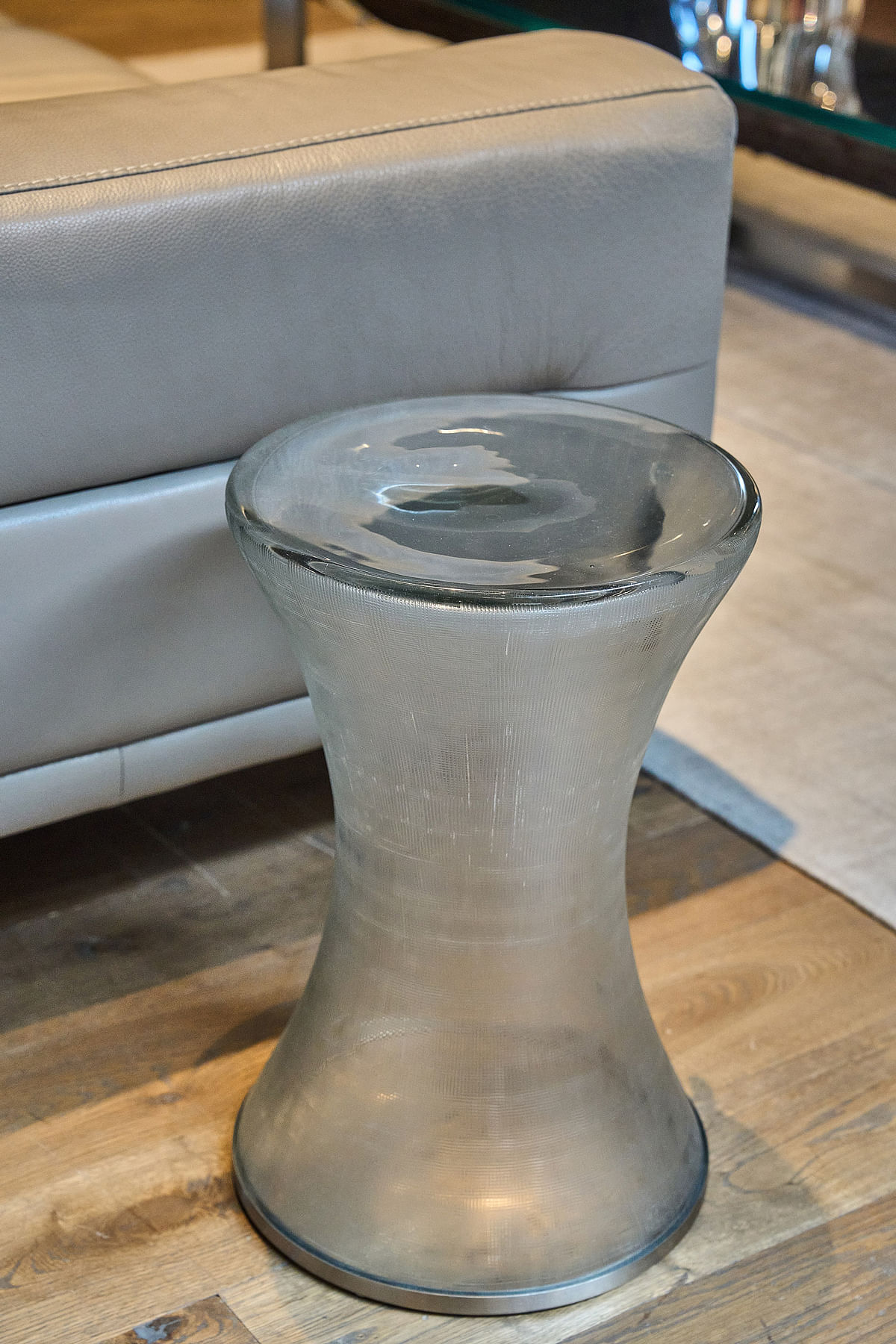 An hourglass-shaped glass side table stands on a wooden floor in front of a grey leather sofa. 