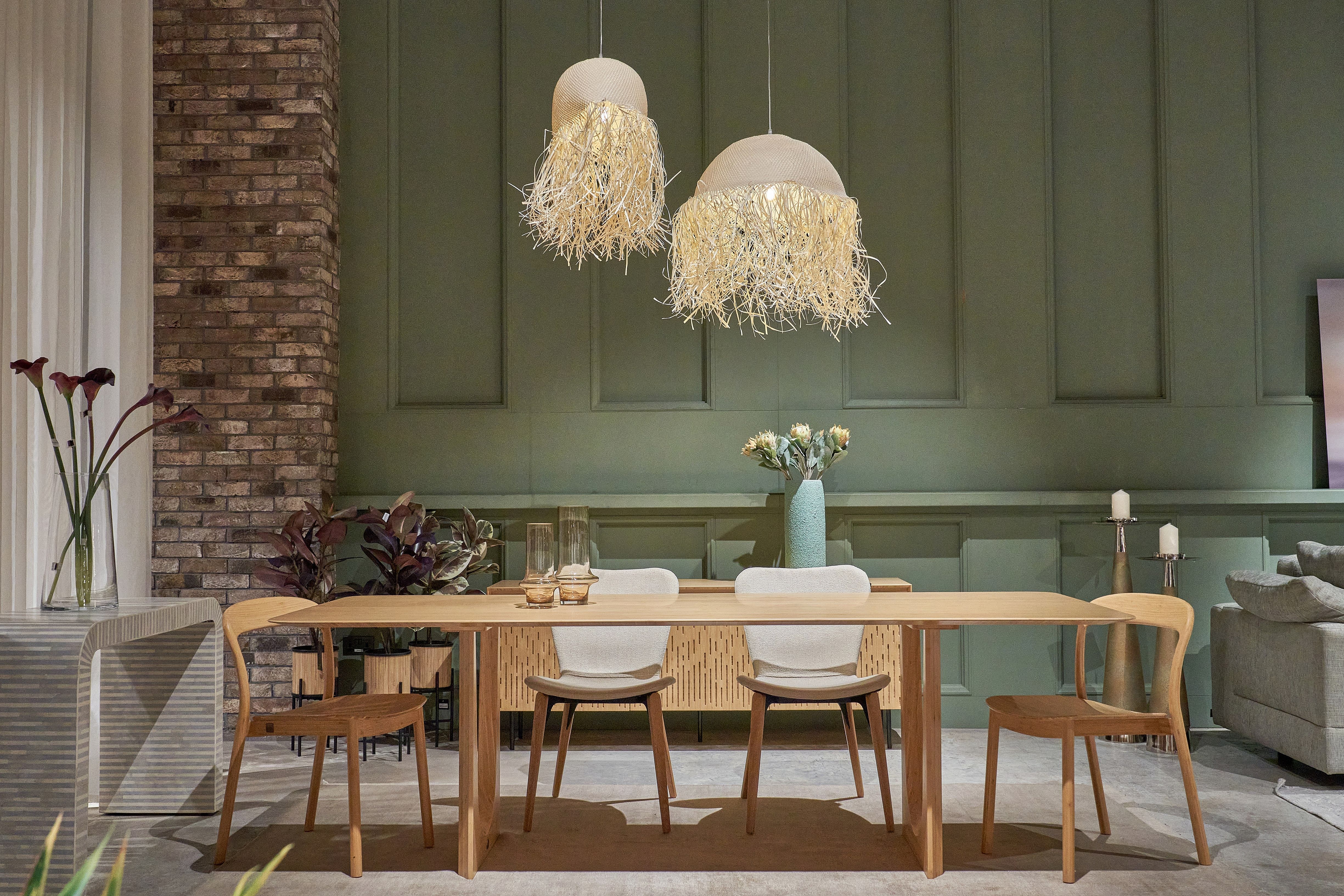 Palm leaf pendant lights hang above a wooden dining table and dining chairs set against a green and exposed brick wall.