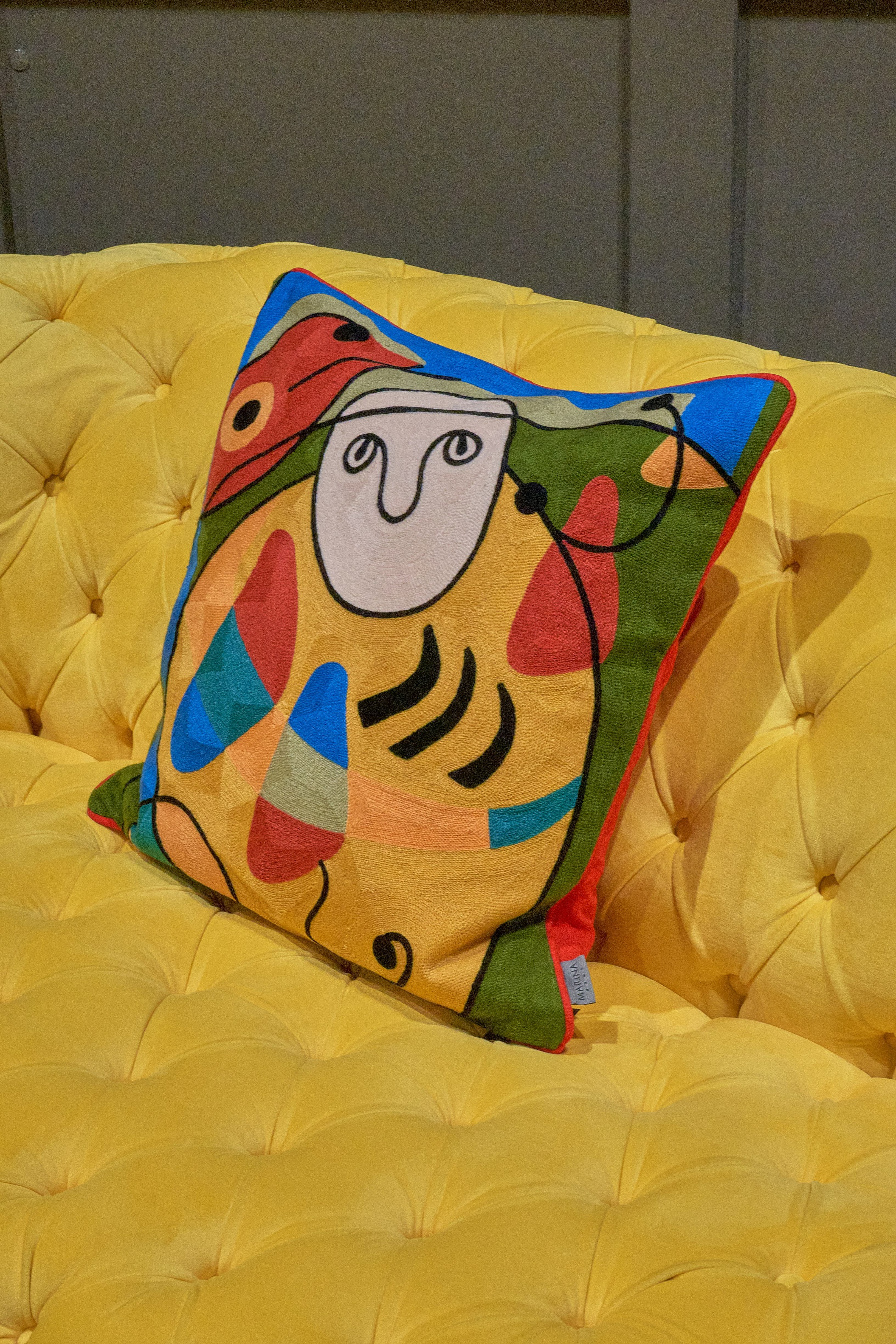A square cushion with an abstract face and patterns in vibrant blues, greens, and oranges sits on a yellow sofa.