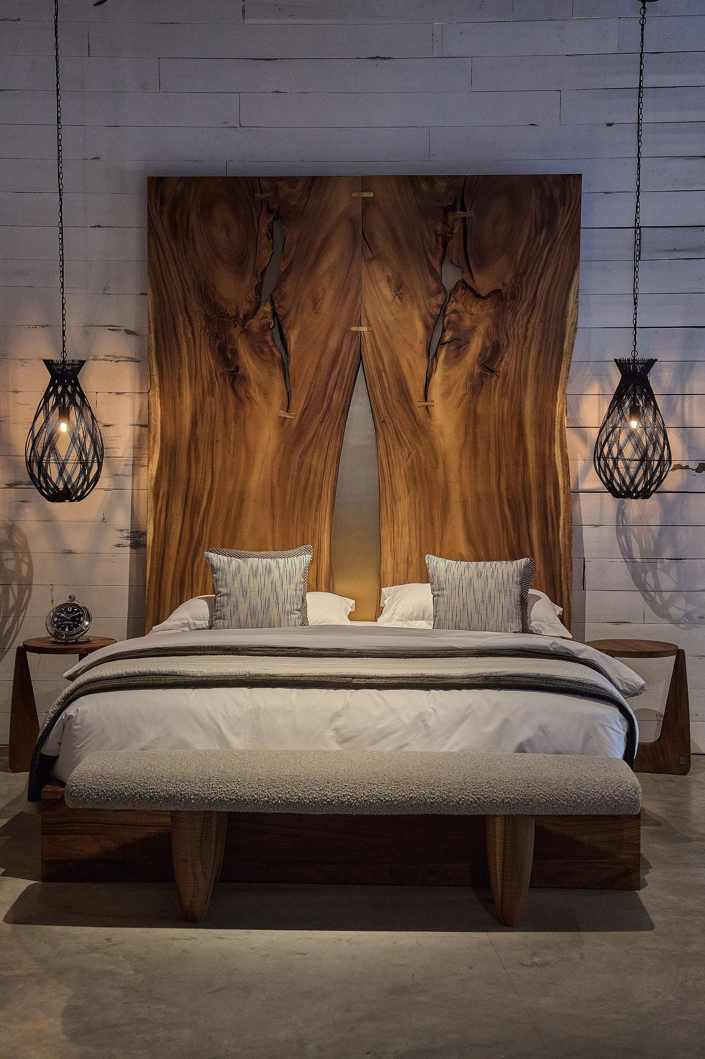 A bed with a huge wooden headboard sits alongside two pendant lights hanging from the ceiling, with a bench in front.