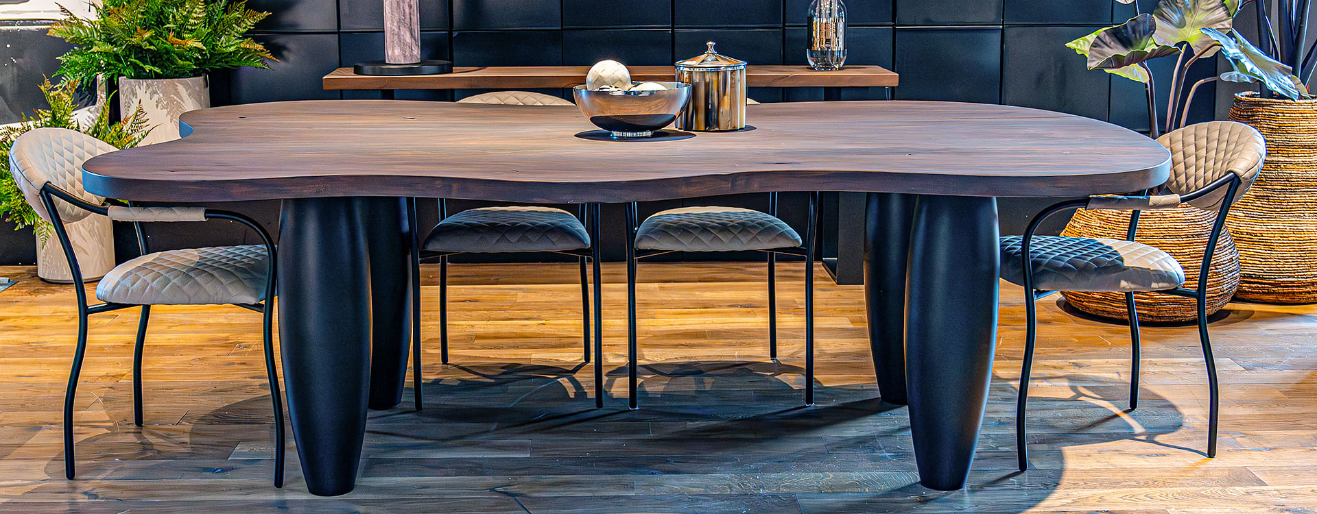 A round-edged dining table topped with silverware sits alongside quilted chairs on a wooden floor. Potted plants stand to the side, and a console table to the back holds a lamp and vase of flowers. 