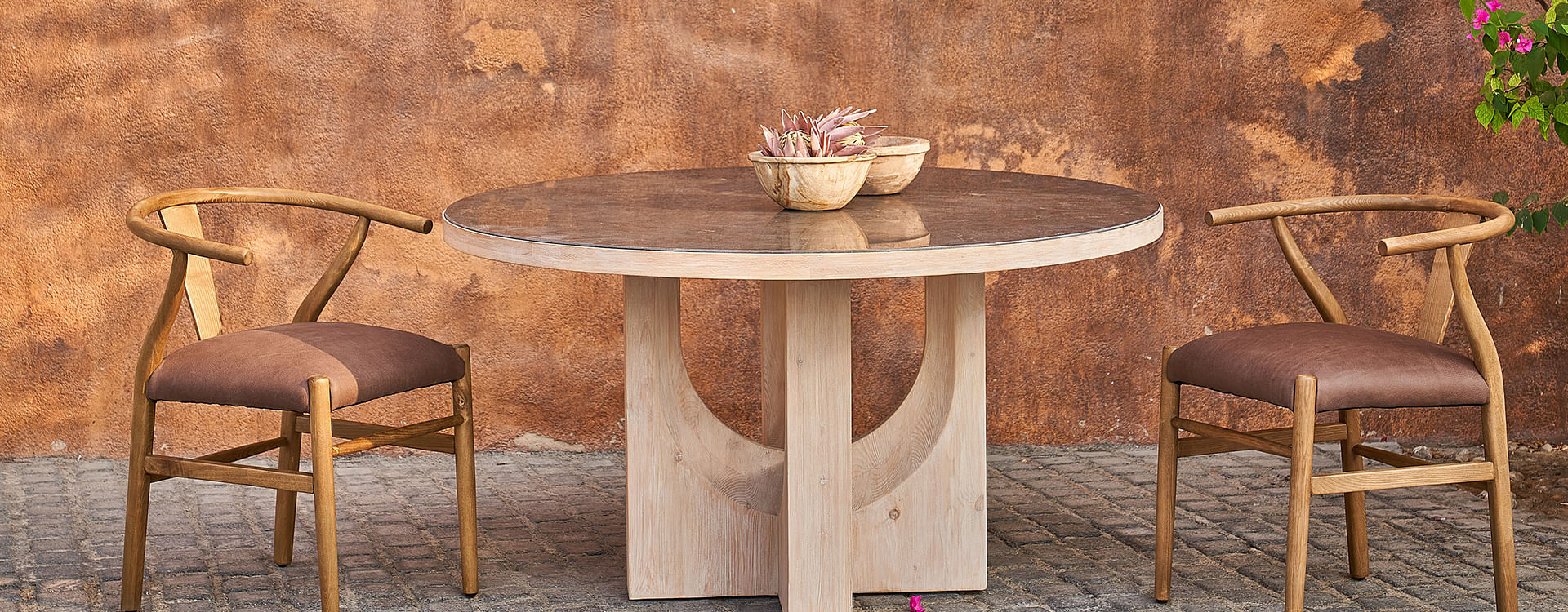 A light brown dining table and leather dining chairs sit on a brick floor against a brown wall.