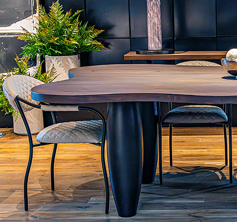 A round-edged dining table topped with silverware sits alongside quilted chairs on a wooden floor. Potted plants stand to the side, and a console table to the back holds a lamp and vase of flowers. 