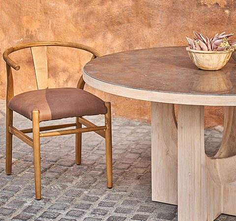 A light brown dining table and leather dining chairs sit on a brick floor against a brown wall.