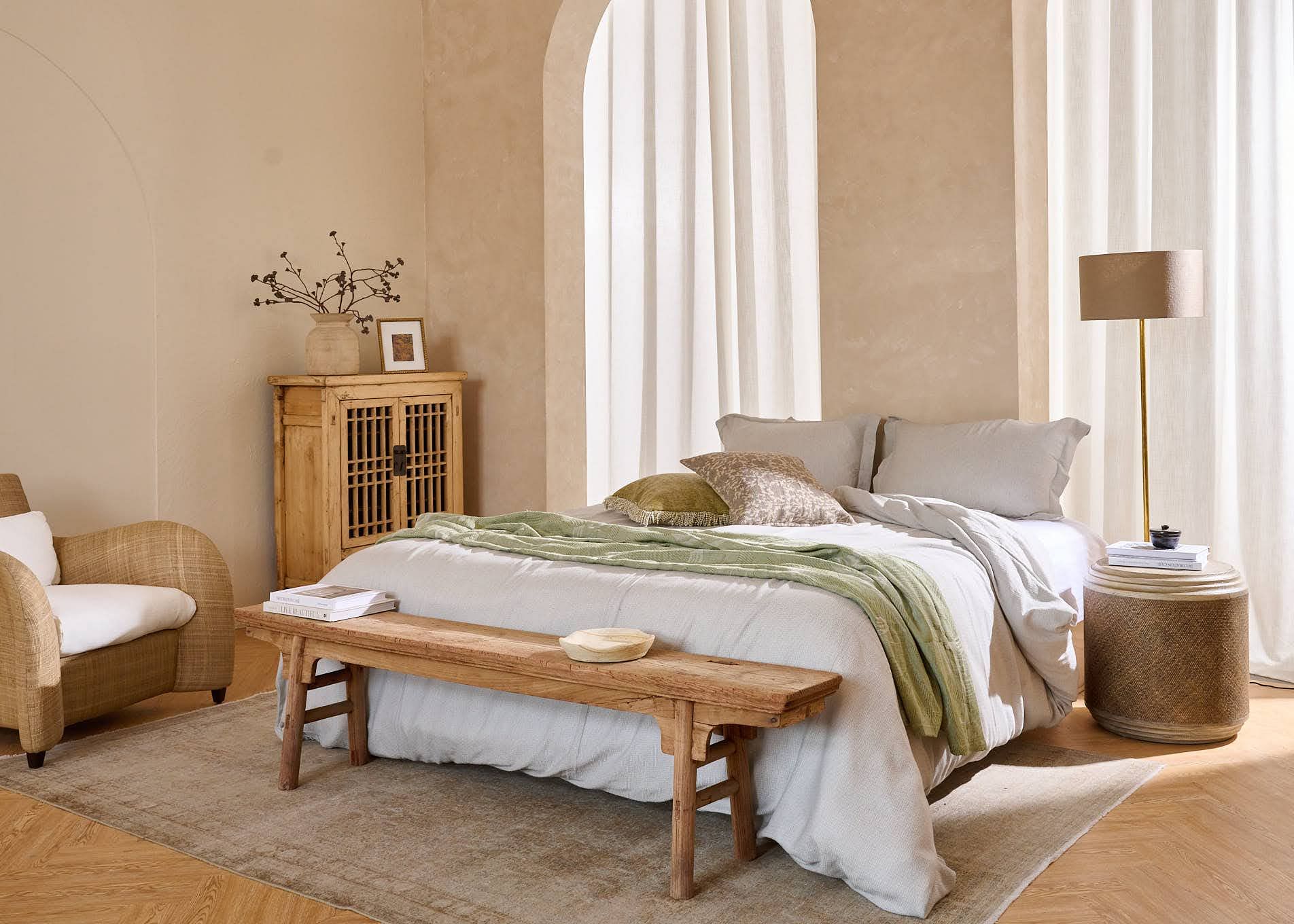 A bed is made up with bed linens. A wooden bench stands to the front, with a standing lamp and side table to one side. 